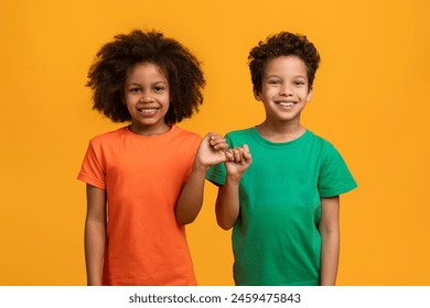 African American young boy and girl are standing side by side, holding hands with bright smiles on their faces, both wearing colorful t-shirts, standing against a vivid yellow backdrop - Powered by Shutterstock