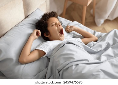 African American young boy is laying in bed, his mouth wide open in a yawn. He appears tired as he stretches his arms out. The background shows a cozy bedroom setting. - Powered by Shutterstock