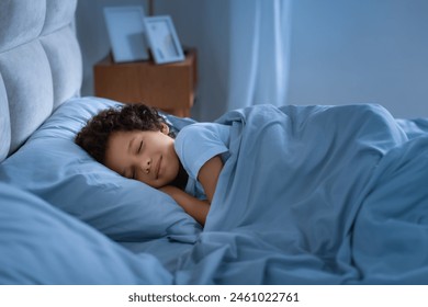 African American young boy peacefully sleeps in a bed with blue sheets, his eyes closed in a state of relaxation. The room is dimly lit with a warm glow, creating a cozy atmosphere - Powered by Shutterstock