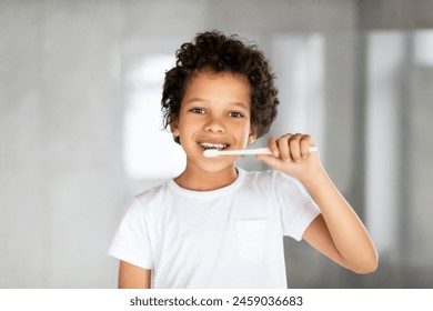 African American young boy with short hair is diligently brushing his teeth with a blue toothbrush in front of a mirror. He is standing in a bathroom, focused on his oral hygiene routine. - Powered by Shutterstock