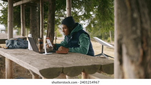 African American young boy sits in wooden gazebo, talks at video call using laptop and phone on tripod. Teenager remotely studies during holidays in beautiful mountain forest. Outdoor study concept. - Powered by Shutterstock