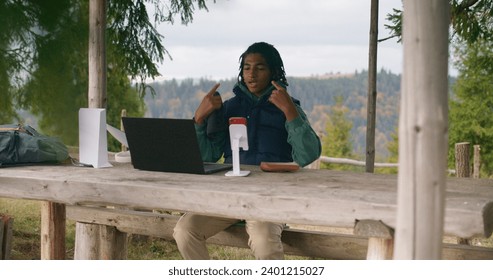 African American young boy sits in wooden gazebo and talks at video call using laptop and phone on tripod. Teenager remotely studies during holidays in mountain forest. Concept of outdoor studying. - Powered by Shutterstock