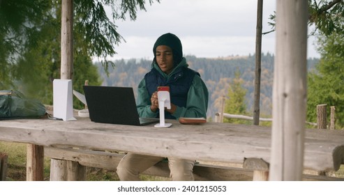 African American young boy sits in wooden gazebo and talks at video call using laptop and phone on tripod. Teenager remotely studies during holidays in mountain forest. Concept of outdoor studying. - Powered by Shutterstock