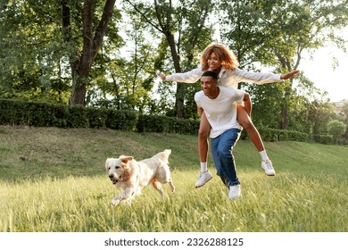 african american young couple in love runs with dog in the park in the summer and smiles, the guy carries his girlfriend on his back and walks with golden retriever outdoors on the grass - Powered by Shutterstock