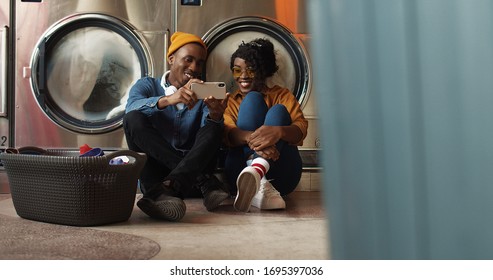 African American young couple sitting on floor with basket of dirty clothes and watching video on smartphone while washing machines working Man and woman using phone and resting in public laundromat