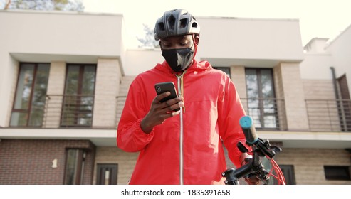 African American Young Deliveryman In Casque And Medical Protective Mask Standing At His Bicycle At Street, Looking Address. Male Courier Using Navigation On Phone To Find Address.