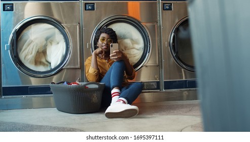 African American young girl sitting on floor with basket of dirty clothes and tapping on smartphone while washing machines working. Women watching video on phone or chatting in public laundromat.  - Powered by Shutterstock