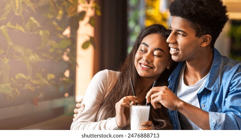 African American young guy and girl are seated at a table in a cafe, both holding milkshakes and sipping through straws. They appear relaxed and engaged in conversation, copy space - Powered by Shutterstock