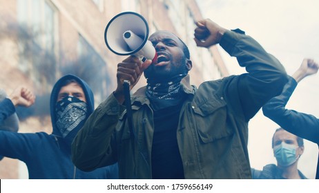 African American young handsome guy with scarf on face protesting in middle of crowd of protesters and screaming mottos in megaphone. Guy leading at manifestation and riot for human rights in smoke. - Shutterstock ID 1759656149
