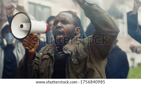African American young handsome man screaming in megaphone at protest for human rights outdoors in smoke. Group of people protesting at street. Strike against violence.