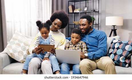 African American young happy nice family with children resting at home on couch spending time together and using digital gadgets. Little girl tapping on tablet while teen boy texting on laptop - Powered by Shutterstock