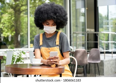 African American Young Hipster Gen Z Young Woman With Afro Hair Wearing Face Mask Holding Smart Phone Using Mobile Apps Sitting At Table In Outdoor Cafe. Social Distancing Everyday Life Tech Concept