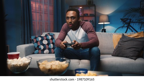 African American young joyful man resting at home sitting on couch and playing video games in evening alone. Handsome guy gamer plays game on console having fun at night. Entertainment concept - Powered by Shutterstock