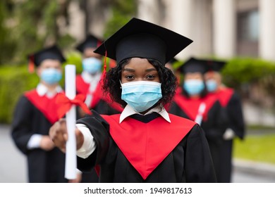 African American Young Lady Student In Graduation Costume And Protective Face Mask Posing Over International Group Of Students At University Campus, Showing Her Diploma, 2021 Graduation Concept