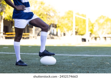 African American young male athlete standing on a rugby ball on field outdoors, copy space. He has dark skin, wearing a blue jersey, white shorts, and rugby cleats, unaltered. - Powered by Shutterstock