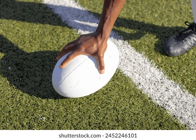 An African American young male athlete wearing cleats placing hand on rugby ball on grass field. Sunlight casting shadows on green turf, marking the start of a game, unaltered. - Powered by Shutterstock