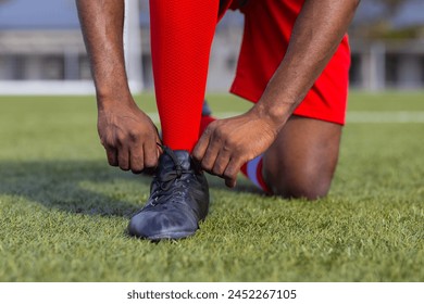 African American young male athlete tying shoelaces on soccer field outdoors. Wearing red shorts, black shoes, and showing strong legs, unaltered. - Powered by Shutterstock