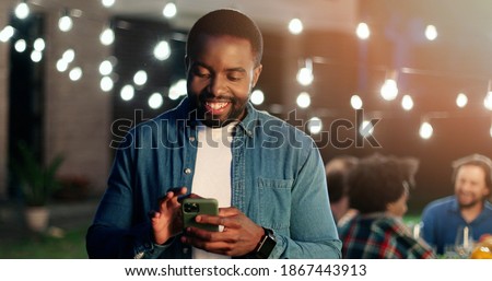 African American young man standing outdoor at night party, smiling and using smartphone. Lights on background. Guy tapping and scrolling on mobile phone and laughing. Texting message. Chatting.