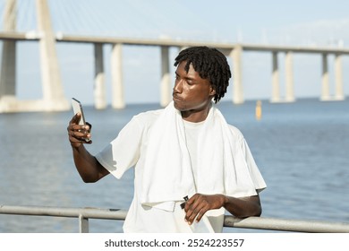 African American young man stands by the water, holding his phone to take a selfie. He wears a white shirt and a towel around his neck, with a bridge visible behind him on a clear day. - Powered by Shutterstock