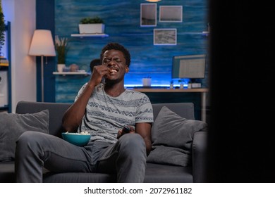 African American Young Man Watching Television Laughing During Comedy Movie Relaxing In Living Room. Black Guy Sitting On Sofa While Eating Popcorn Enjoying Spending Tine Alone