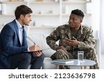 African american young military man in camouflage outfit having conversation with guy in suit social , sitting on couch at home, black soldier attending therapy session with psychologist