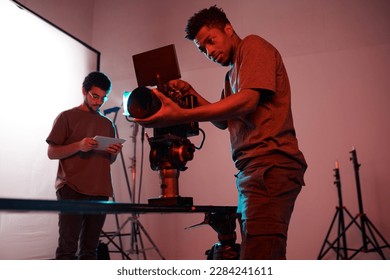 African American young operator setting his camera before shooting with his colleague using tblet pc in background - Powered by Shutterstock