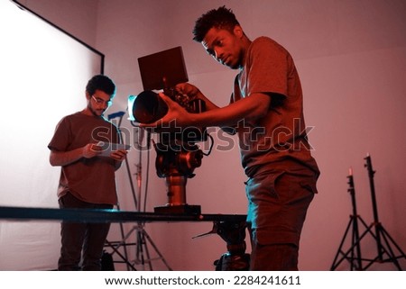 African American young operator setting his camera before shooting with his colleague using tblet pc in background