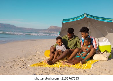 African American Young Parents With Children Sitting Under Umbrella On Blanket At Beach Against Sky. Copy Space, Nature, Unaltered, Childhood, Family, Togetherness, Summer, Enjoyment, Holiday.