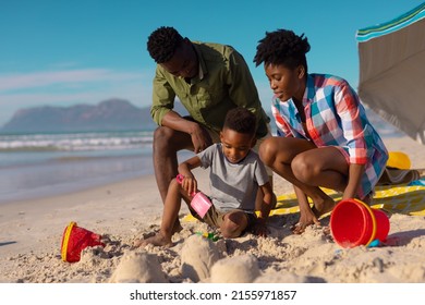 African American Young Parents Looking At Son Playing With Sand At Beach Against Sky On Sunny Day. Summer, Nature, Unaltered, Beach, Childhood, Family, Togetherness, Lifestyle, Enjoyment, Holiday.