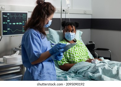 African American Young Patient With Protection Face Mask Against Covid19 Lying In Bed Explaining Sickness Symptoms To Medical Nurse In Hospital Office. Assistant Checking Healthcare Treatment