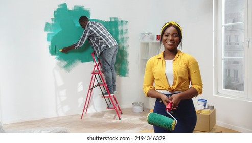 African American young pretty woman stands in room looking to camera and smiling, on the background husband painting wall in green color with roller brush - Powered by Shutterstock