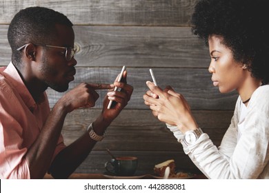 African American young students sitting at a cafe table using mobile phones, texting via social networks with obsessed expression, not looking and talking to each other. Modern technology addiction