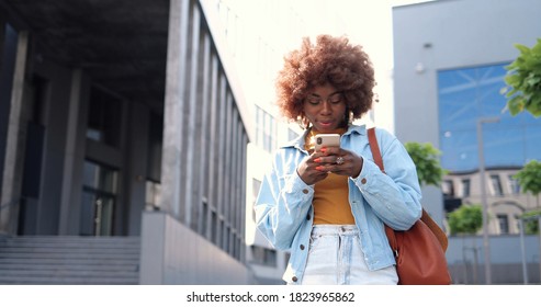 African American Young Stylish Curly Woman Tapping Or Scrolling On Smartphone And Standing At City Street. Beautiful Female Texting Message On Mobile Phone And Chatting. Outside.