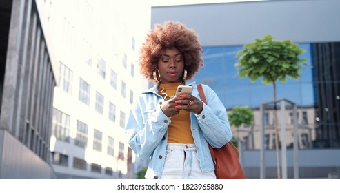 African American Young Stylish Curly Woman Tapping Or Scrolling On Smartphone And Standing At City Street. Beautiful Female Texting Message On Mobile Phone And Chatting. Outside.