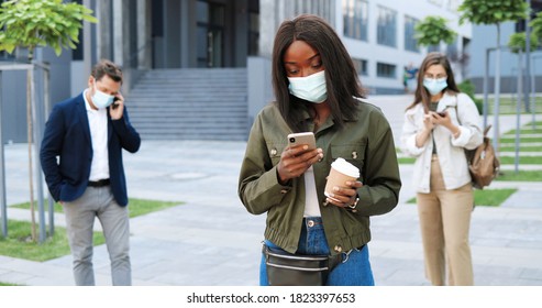 African American young stylish woman in medical mask standing at street and texting message on smartphone. Female outdoor tapping on mobile phone and holding coffee. Pandemic. Mixed-races people. - Powered by Shutterstock