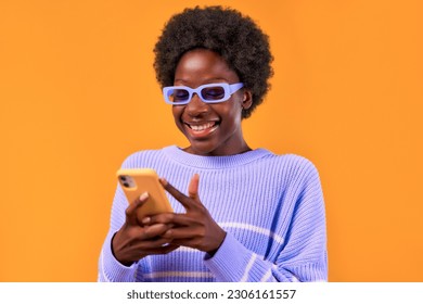 African American young woman with afro hair styling standing in a blue sweater and glasses on a bright orange background using the phone and smiling cheerfully. - Powered by Shutterstock