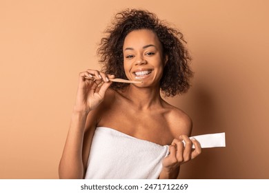 African American young woman with curly hair smiles brightly while brushing her teeth with a bamboo toothbrush. She is wrapped in a white towel and holds a tube of toothpaste in her other hand - Powered by Shutterstock