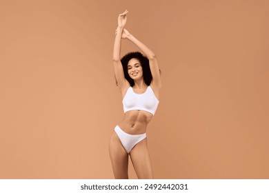 African American young woman with long, dark hair is wearing a white sports bra and matching underwear. She is standing with her arms raised above her head and is smiling at the camera. - Powered by Shutterstock