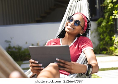 African American young woman relaxing in hammock at home, holding tablet. She has braided hair, sunglasses, and casual clothes, enjoying sunny day, unaltered - Powered by Shutterstock