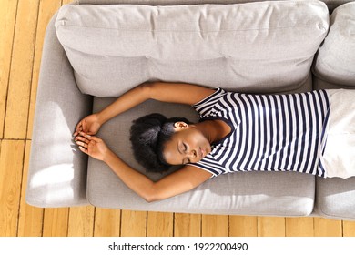 African American Young Woman Wear Stripped T-shirt Sleeping On Couch With Arms Up At Home, Closing Eyes And Taking A Break. Black Millennial Girl Resting On Sofa, Top View. Relax, Doze, Lazy Day.