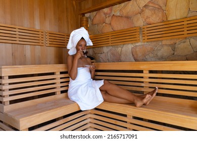 African american young woman wearing bathrobe drinking tea while sitting on wooden bench at sauna. unaltered, spa, home, relaxation, drink, pampering, detox, body care and wellbeing concept.