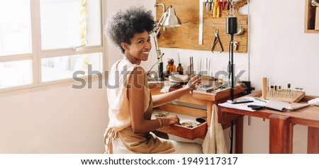 African artist glancing back while working at jewelry workshop. Cheerful young female worker in a jewelry workshop.