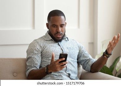 African astonished man sitting on sofa holds cellphone read e-mail sms feels shocked received terrified news, guy looks at online calendar forgot missed important meeting, phone crash problems concept