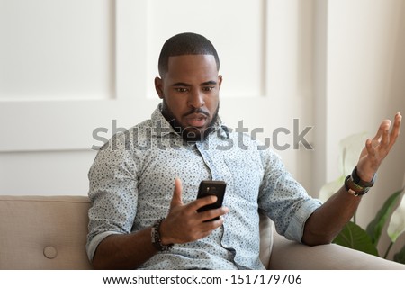 African astonished man sitting on sofa holds cellphone read e-mail sms feels shocked received terrified news, guy looks at online calendar forgot missed important meeting, phone crash problems concept