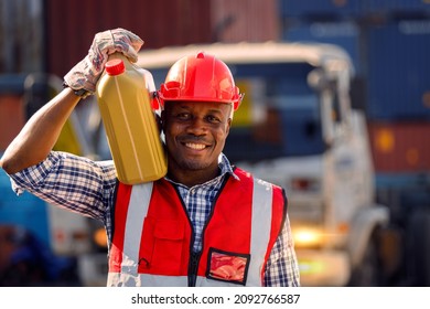 An African Automotive Mechanic Engineer In Protective Clothing And A Hard Hat Stands Holding Motor Engine Oil To Transfer To A Transportation Truck. Maintenance Man In The Automotive Industry.