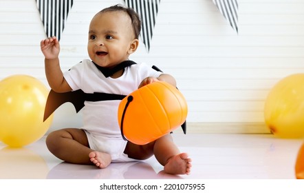 African Baby Kid Dressing Up In Vampire Fancy Halloween Costume With Black Bat Wings, Little Cute Child Go To Party, Playing Trick Or Treat, Holding Orang Pumpkin Basket. Happy Halloween Celebration.