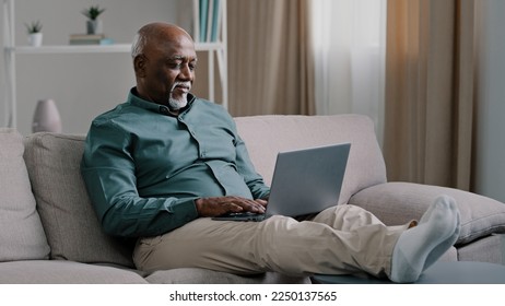 African bald overweight obese old middle-aged man senior businessman sitting on sofa at home working with laptop typing feels tired stress relaxing leaning back on sofa pauses after finishing work - Shutterstock ID 2250137565