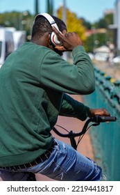 African Black Boy Listening To Music With The Headphones On The Bike. Urban And Street Scenery On A Bridge