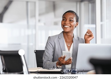 African black business woman using smartphone while working on laptop at office. Smiling mature african american businesswoman looking up while working on phone. Successful woman entrepreneur. - Powered by Shutterstock