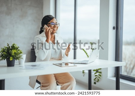 African black business woman using smartphone while working on laptop at office. Smiling african american businesswoman lookingup at copy space while working on phone. 